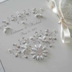 May include: A white wedding guest book with a gray floral design and pearl embellishments. The book is tied with a white satin ribbon and the text "Catherine & Michael May 15, 2015" is written in gray ink.