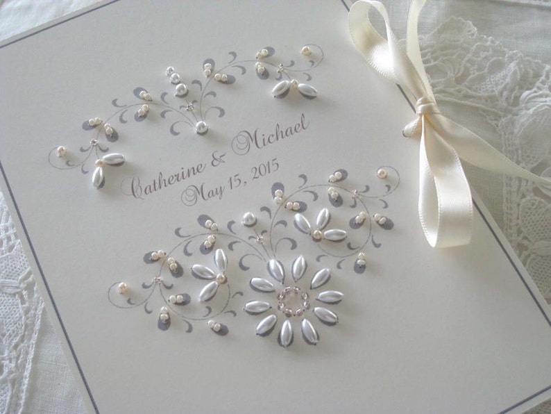 May include: A white wedding guest book with a gray floral design and pearl embellishments. The book is tied with a white satin ribbon and the text "Catherine & Michael May 15, 2015" is written in gray ink.