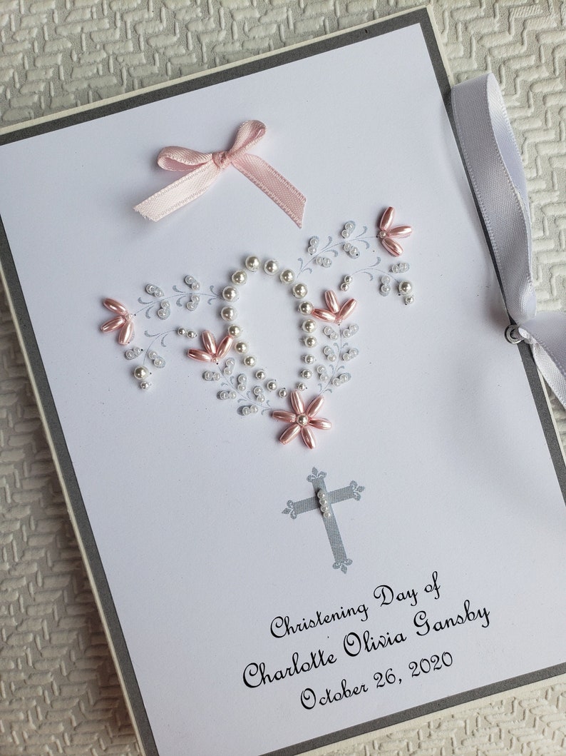 May include: A white card with a gray border and a pink bow. The card is decorated with white and pink beads in the shape of a wreath. The text on the card reads "Christening Day of Charlotte Olivia Gansby October 26, 2020".