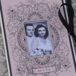 May include: A pink wedding album with an ornate, black and white design. The album features a photo of a couple in a black and white photo, with the text "John & Heidi" on the front.