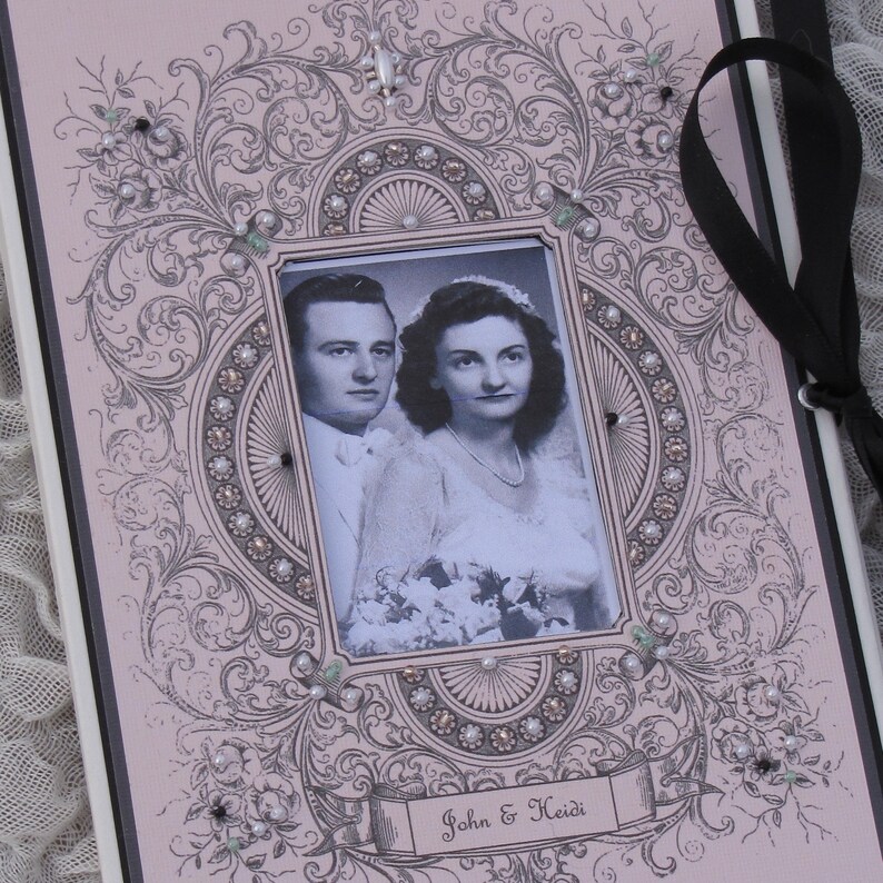 May include: A pink wedding album with an ornate, black and white design. The album features a photo of a couple in a black and white photo, with the text "John & Heidi" on the front.