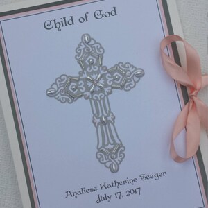 May include: A white card with a gray cross decorated with silver beads and the text "Child of God" at the top. The card also includes the name "Analiese Katherine Seeger" and the date "July 17, 2017".