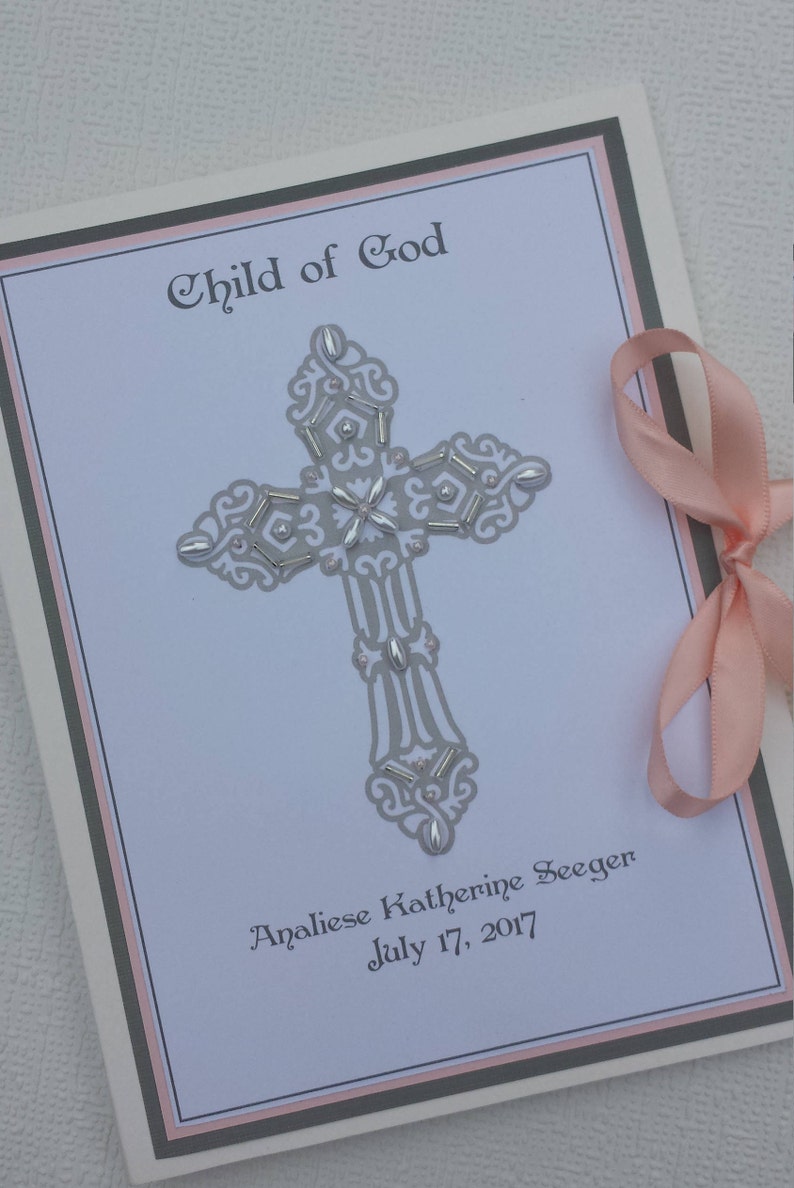 May include: A white card with a gray cross decorated with silver beads and the text "Child of God" at the top. The card also includes the name "Analiese Katherine Seeger" and the date "July 17, 2017".