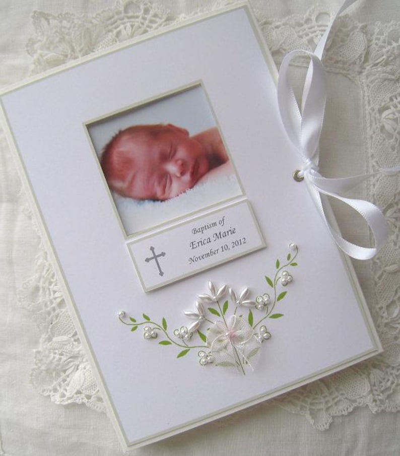 May include: White photo album with a white satin ribbon closure. The album has a photo frame with a picture of a baby. The album has a silver cross and the text "Baptism of Erica Marie November 10, 2012".