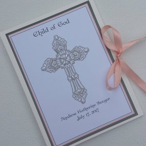 May include: A white card with a silver cross and the text "Child of God" in a cursive font. The card also includes the name "Analiese Katherine Seeger" and the date "July 17, 2017". The card is decorated with a pink ribbon.