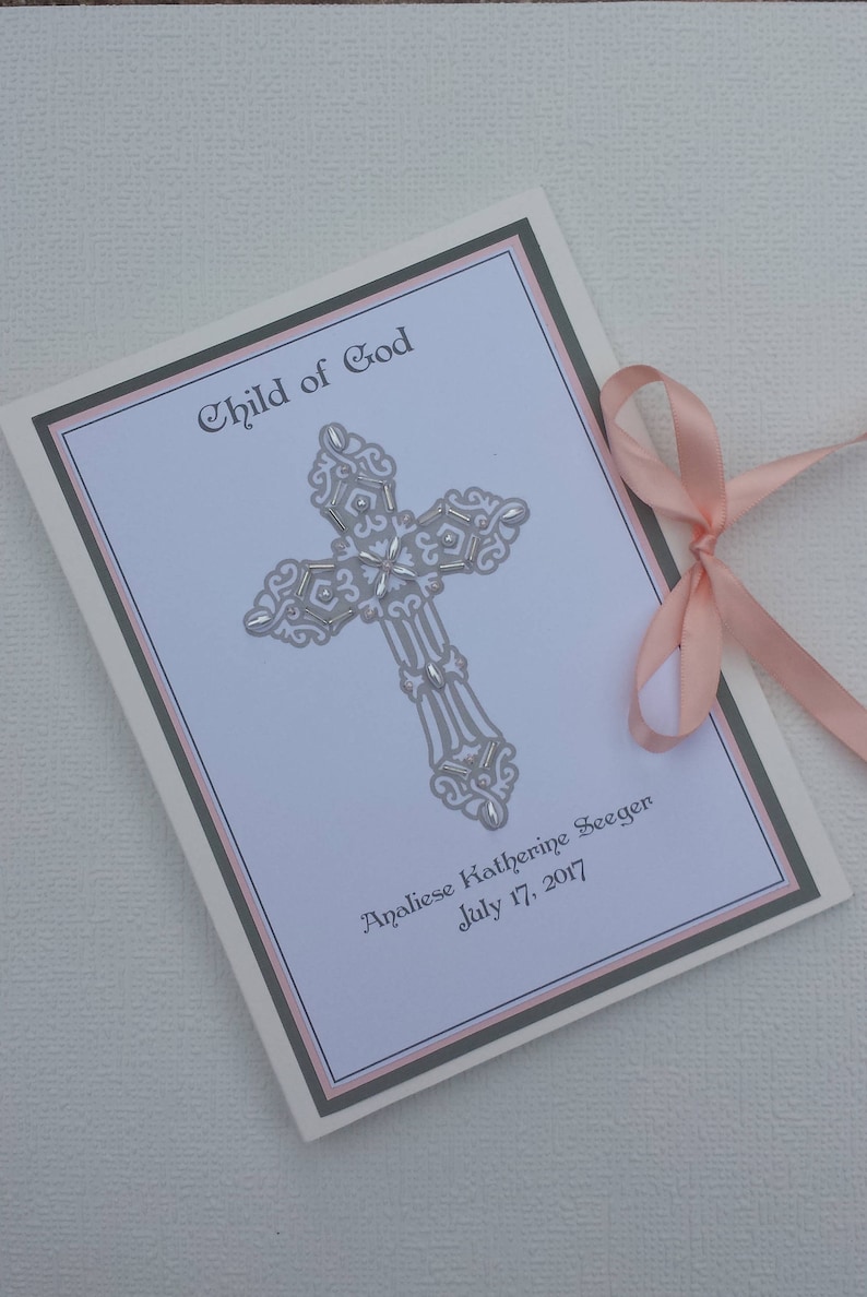 May include: A white card with a silver cross and the text "Child of God" in a cursive font. The card also includes the name "Analiese Katherine Seeger" and the date "July 17, 2017". The card is decorated with a pink ribbon.