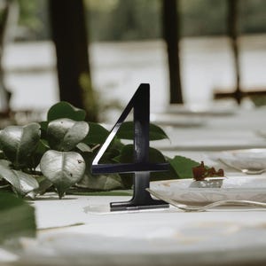 Puede incluir: Un número de mesa negro, el número cuatro, hecho de acrílico, sobre un mantel blanco. El número está frente a una guirnalda de hojas verdes y platos blancos. El fondo borroso muestra árboles y un cenador.