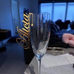 May include: A clear champagne flute with a gold-colored acrylic name tag that reads "Alicia" in an elegant script. The tag is designed to hook onto the rim of the glass, adding a personalized touch to the drinkware.