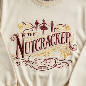 Puede incluir: Una sudadera de color crema con un diseño bordado en color burdeos que representa a una bailarina, dos figuras y el texto "The Nutcracker" con un contorno dorado y el texto "EST. 1903".