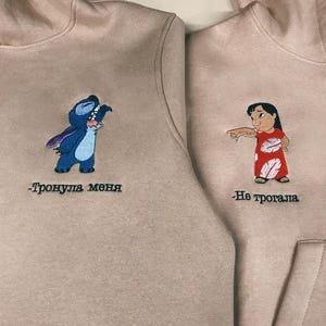Może przedstawiać: Dwie beżowe bluzy z kapturem z haftowanymi postaciami z kresk&oacute;wek. Lewa bluza przedstawia Stitch z Lilo & Stitch z tekstem "Тронула меня", a prawa bluza przedstawia Lilo z Lilo & Stitch z tekstem "Не трогала".