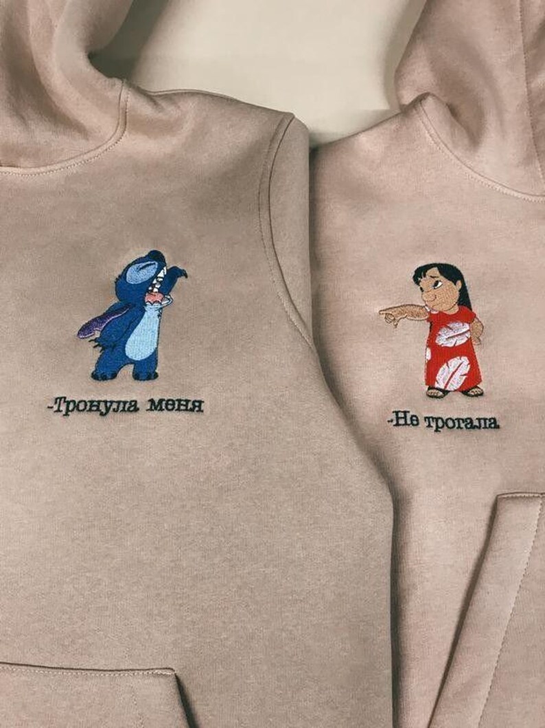Może przedstawiać: Dwie beżowe bluzy z kapturem z haftowanymi postaciami z kresk&oacute;wek. Lewa bluza przedstawia Stitch z Lilo & Stitch z tekstem "Тронула меня", a prawa bluza przedstawia Lilo z Lilo & Stitch z tekstem "Не трогала".