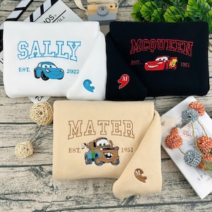 May include: Three folded sweatshirts with embroidered designs. One is white with "SALLY" and a blue car, another is black with "MCQUEEN" and a red car, and the third is tan with "MATER" and a brown car. Each has a matching heart detail.