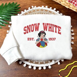 May include: White sweatshirt with the words "SNOW WHITE" in red, collegiate-style lettering, and an illustration of Snow White holding an apple. The sweatshirt also features "EST. 1937".