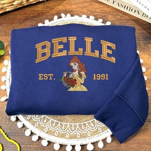 May include: A navy blue sweatshirt with gold embroidered text reading "BELLE" above an illustration of a woman in a yellow dress holding flowers. The text "EST. 1991" is below. The sweatshirt has a ribbed cuff in navy blue.