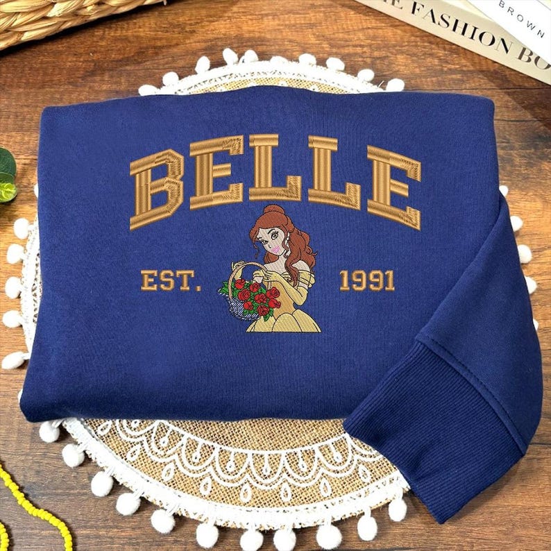 May include: A navy blue sweatshirt with gold embroidered text reading "BELLE" above an illustration of a woman in a yellow dress holding flowers. The text "EST. 1991" is below. The sweatshirt has a ribbed cuff in navy blue.