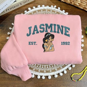 May include: A pale pink sweatshirt with the name "JASMINE" in teal lettering and an illustration of Princess Jasmine. Below the illustration are the words "EST. 1992". The sweatshirt has a ribbed cuff and a round neckline.