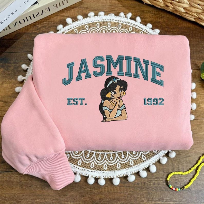 May include: A pale pink sweatshirt with the name "JASMINE" in teal lettering and an illustration of Princess Jasmine. Below the illustration are the words "EST. 1992". The sweatshirt has a ribbed cuff and a round neckline.