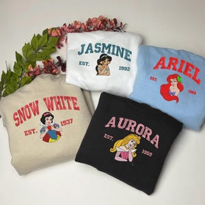 May include: Five sweatshirts with Disney princess designs. The sweatshirts are in white, light blue, black, and beige. Each sweatshirt has a princess's name, a graphic of the princess, and the year of the movie's release.