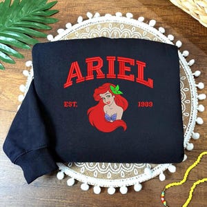 May include: Black sweatshirt featuring the name "ARIEL" in red embroidered letters. Below the name is an image of Ariel with red hair and a green leaf. The text "EST. 1989" is also embroidered on the sweatshirt.