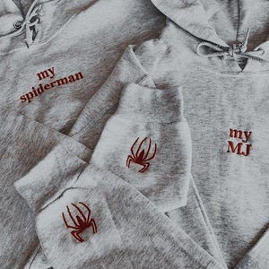 Puede incluir: Dos sudaderas con capucha grises con dise&ntilde;os de ara&ntilde;as bordadas en rojo y texto. Una sudadera dice "my spiderman" y la otra dice "my MJ".