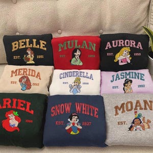 May include: A selection of sweatshirts with embroidered Disney princess designs. Each sweatshirt features a princess's name, a graphic of the character, and the year of establishment. Colours include black, red, beige, white, pink, green, navy, and grey.