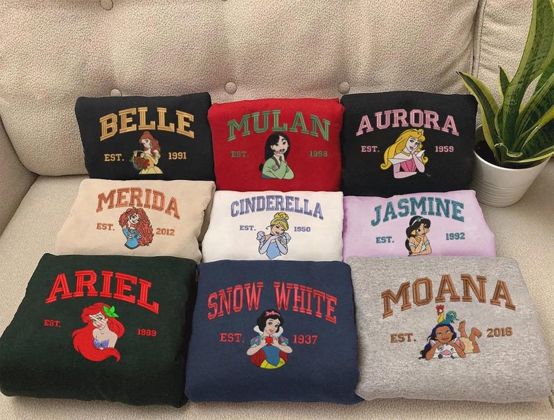 May include: A selection of sweatshirts with embroidered Disney princess designs. Each sweatshirt features a princess's name, a graphic of the character, and the year of establishment. Colours include black, red, beige, white, pink, green, navy, and grey.
