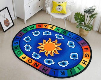 May include: Oval blue rug with a colorful alphabet border and a central design featuring a sun and clouds. The rug also has numbers and is designed for educational purposes. The rug is on a light-colored floor.
