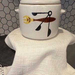 May include: A white ceramic kitchen storage jar with a lid, decorated with a brown and yellow utensil design. It rests on a white fringed cloth. The background has a hexagonal tile pattern.