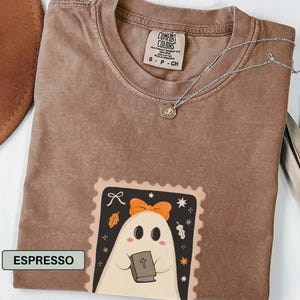K&ouml;nnte beinhalten: Espressofarbenes T-Shirt mit einem Geister-Motiv, das ein Buch h&auml;lt. Der Geist hat eine orangefarbene Schleife und befindet sich in einem dekorativen Rahmen. Eine silberne Halskette mit Anh&auml;nger befindet sich auf dem Shirt.