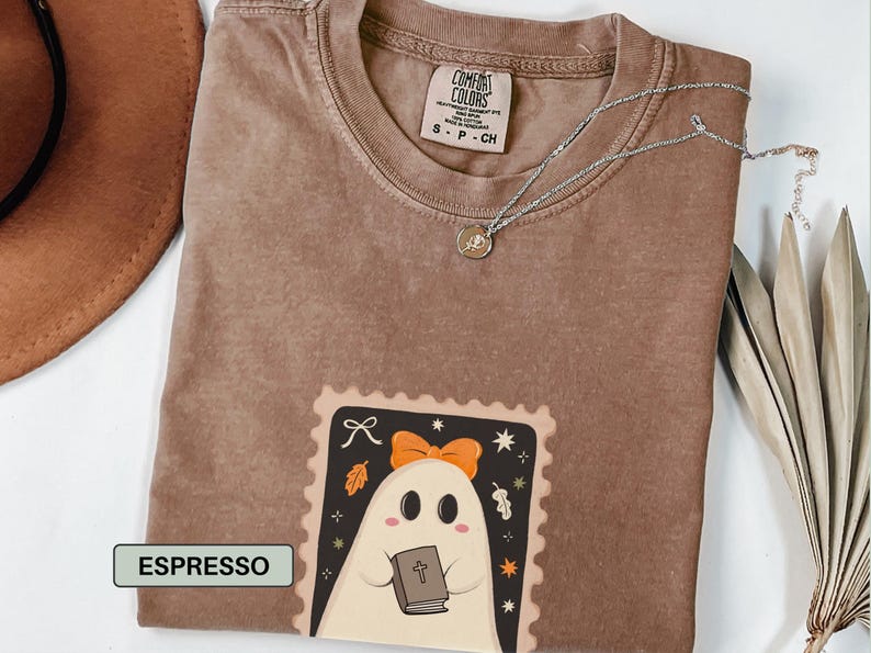 K&ouml;nnte beinhalten: Espressofarbenes T-Shirt mit einem Geister-Motiv, das ein Buch h&auml;lt. Der Geist hat eine orangefarbene Schleife und befindet sich in einem dekorativen Rahmen. Eine silberne Halskette mit Anh&auml;nger befindet sich auf dem Shirt.