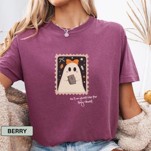 K&ouml;nnte beinhalten: Beerenfarbenes T-Shirt mit einem stempelartigen Aufdruck eines Geistes, der ein Buch und eine orangefarbene Schleife h&auml;lt. Der Text auf der Grafik lautet "Ain't no ghost like the Holy Ghost". Es ist ein Freizeitkleidungsst&uuml;ck.