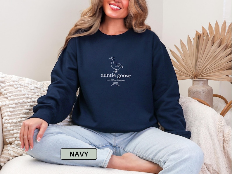 Może przedstawiać: Granatowa bluza z napisem "auntie goose on the loose" i ilustracją gęsi. Bluza jest w jednolitym granatowym kolorze. Słowo "NAVY" znajduje się w prostokącie na dole obrazu.