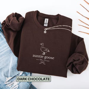 Może przedstawiać: Ciemnobrązowa bluza z napisem "auntie goose on the loose" i grafiką gęsi. Srebrny naszyjnik jest przerzucony przez bluzę. Jasnoniebieskie dżinsy są widoczne po lewej stronie zdjęcia.