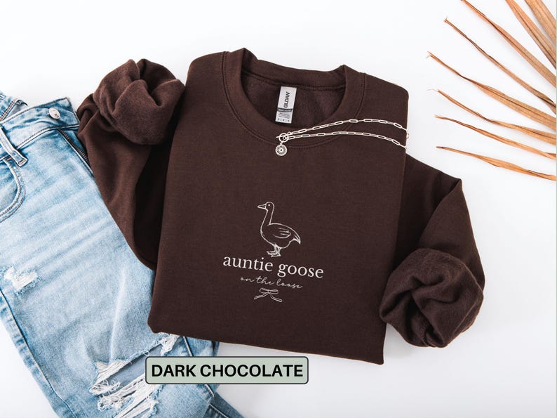 Może przedstawiać: Ciemnobrązowa bluza z napisem "auntie goose on the loose" i grafiką gęsi. Srebrny naszyjnik jest przerzucony przez bluzę. Jasnoniebieskie dżinsy są widoczne po lewej stronie zdjęcia.