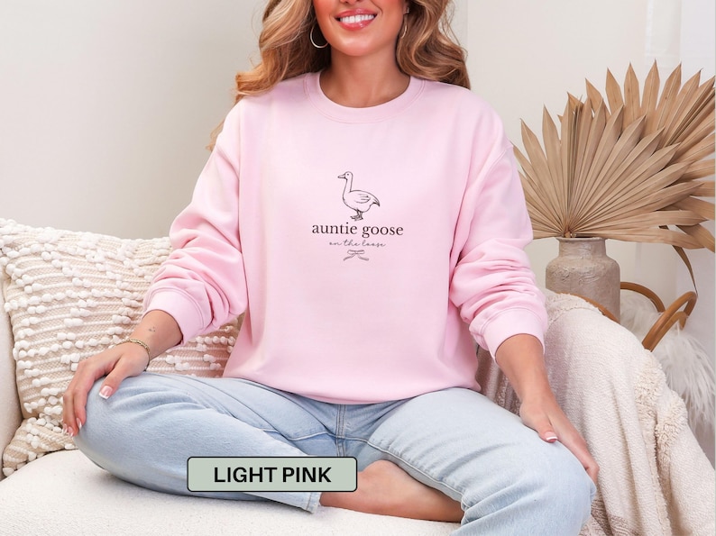 Może przedstawiać: Jasnor&oacute;żowa bluza z napisem "auntie goose on the loose" i ilustracją gęsi. Tekst "light pink" znajduje się w prostokącie na dole obrazu. Bluza jest w kolorze jasnor&oacute;żowym.