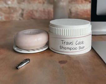 Könnte beinhalten: Ein weißer Reisebehälter mit einem braunen Etikett, auf dem "Travel Case Shampoo Bar" steht. Neben dem Behälter befindet sich eine hellrosa Shampoo-Bar in einer weißen Schale.