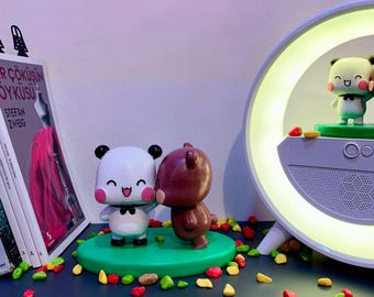 May include: Two small figurines, one white and one brown, are standing on a green base. The white figurine is a panda with a black bow tie and a happy expression. The brown figurine is a bear with a happy expression. The figurines are surrounded by small, colorful stones.