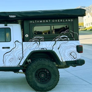 May include: White Jeep Rubicon with black off-road tires and a custom camper top. The camper features the text "ALTAMONT OVERLAND" and a topographic map design. The Jeep is parked on a paved driveway in front of a house.