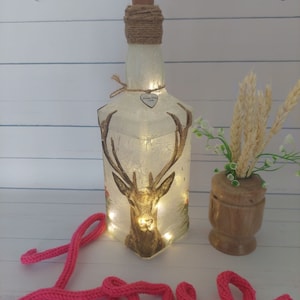 May include: A white glass bottle with a cork stopper and a brown twine bow. The bottle is decorated with a deer image and has a small heart charm with the text "Made with Love". The bottle is lit with white fairy lights and is sitting on a white surface with a pink knitted word "Love".