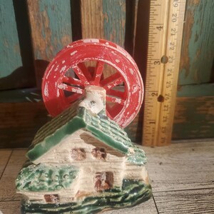 May include: A ceramic water mill figurine with a red water wheel and a green-roofed house. The house has brown window and door details. The water wheel is approximately 2 inches in diameter.