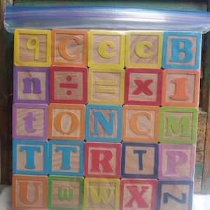 May include: A set of square wooden alphabet and math blocks with colorful letters, numbers, and symbols, arranged in a grid inside a clear plastic storage bag. A clear plastic ruler is positioned at the bottom, showing a length of 6 inches.