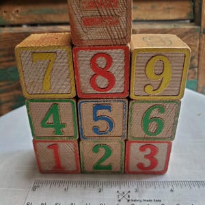 May include: A stack of nine wooden blocks arranged in a three-by-three grid with one block on top, featuring numbers one through nine in various colors. A clear plastic ruler is positioned at the base, showing the width is approximately 4.5 inches.