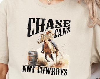 May include: A beige t-shirt featuring a barrel racing design with a woman on a horse. The text "CHASE CANS NOT COWBOYS" is printed in black above and below the image. The shirt is a casual style and suitable for everyday wear.