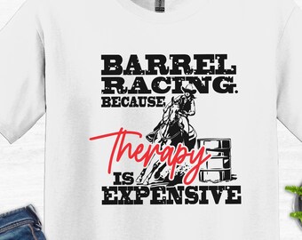 May include: White crew neck t-shirt featuring the text "BARREL RACING. BECAUSE Therapy IS EXPENSIVE" in black and red. The shirt also has a graphic of a horse and rider racing around a barrel. The t-shirt is a casual wear item.