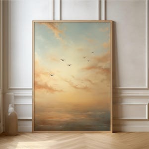 Könnte beinhalten: Ein gerahmter Kunstdruck, der eine ruhige Meereslandschaft mit einem Sonnenuntergangshimmel zeigt. Das Kunstwerk zeigt einen Farbverlauf von sanften Blau- und Rosatönen bis hin zu warmen Orange- und Gelbtönen. Silhouetten von Vögeln sind im Flug.