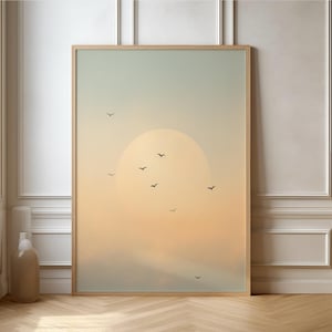 Könnte beinhalten: Ein gerahmter Kunstdruck mit einer ruhigen Sonnenuntergangsszene. Das Kunstwerk zeigt eine große, weiche gelbe Sonne, die hinter einem dunstigen Horizont untergeht, mit mehreren kleinen schwarzen Vögeln im Flug. Der Himmel geht von einem blassen Blau in ein warmes Orange über.