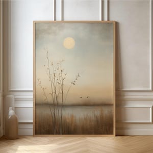 Puede incluir: Una impresión artística enmarcada que presenta una escena serena con un gran sol amarillo pálido en un cielo apagado. Plantas altas y delgadas están en primer plano, con una masa de agua y dos pájaros en vuelo. La obra de arte está en un marco de madera.