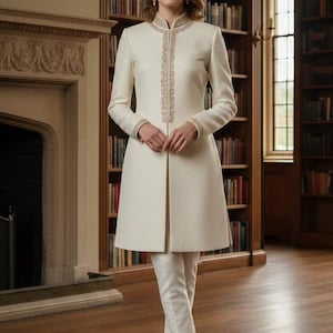 Op de afbeelding: Een crèmekleurige jas en bijpassende broek met gouden versieringen. De jas heeft een hoge kraag en een recht silhouet. De broek is getailleerd. De outfit wordt gedragen door een persoon die in een kamer met een open haard en boekenkasten staat.