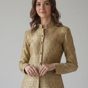 Op de afbeelding: Een goudkleurige jas met lange mouwen, een hoge kraag en een knoopsluiting. De jas heeft een gedetailleerd paisley patroon. Het kledingstuk wordt gedragen door een persoon met bruin haar.