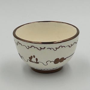May include: A small, round ceramic bowl with an off-white base color. The exterior features hand-painted brown decorative patterns, including wavy lines and floral-like motifs. The rim and the base of the bowl are finished in a solid brown glaze.